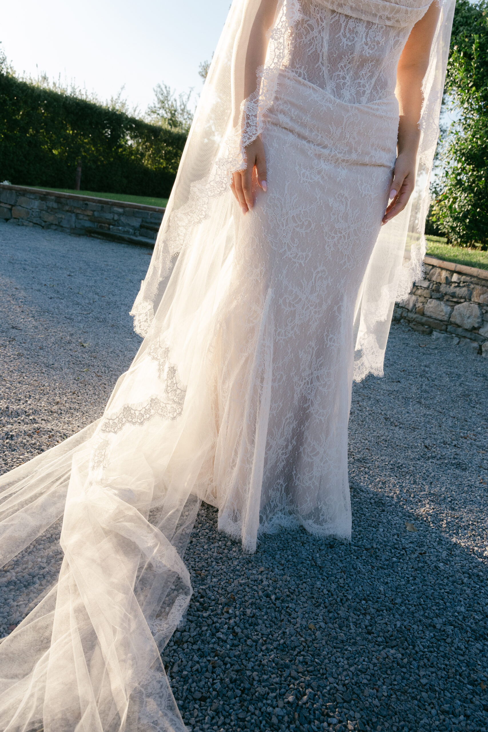 Close detail of bride's lace gown and cathedral veil trailing on a gravel path at Villa Marchese Estate 1795 Tuscany with stone wall behind