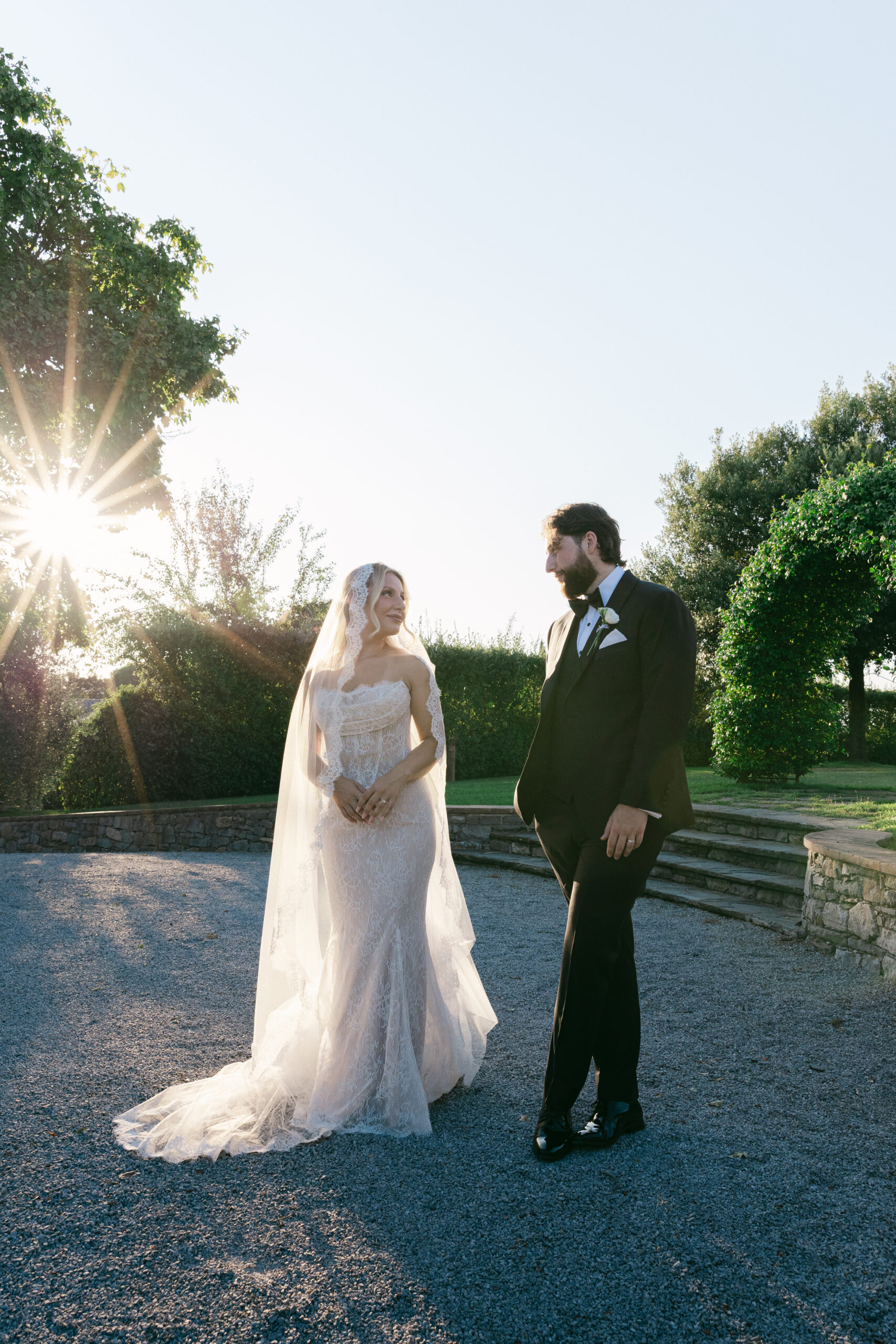 Bride and groom facing each other on a gravel path at Villa Marchese Estate 1795 Tuscany with strong sun flare through the trees