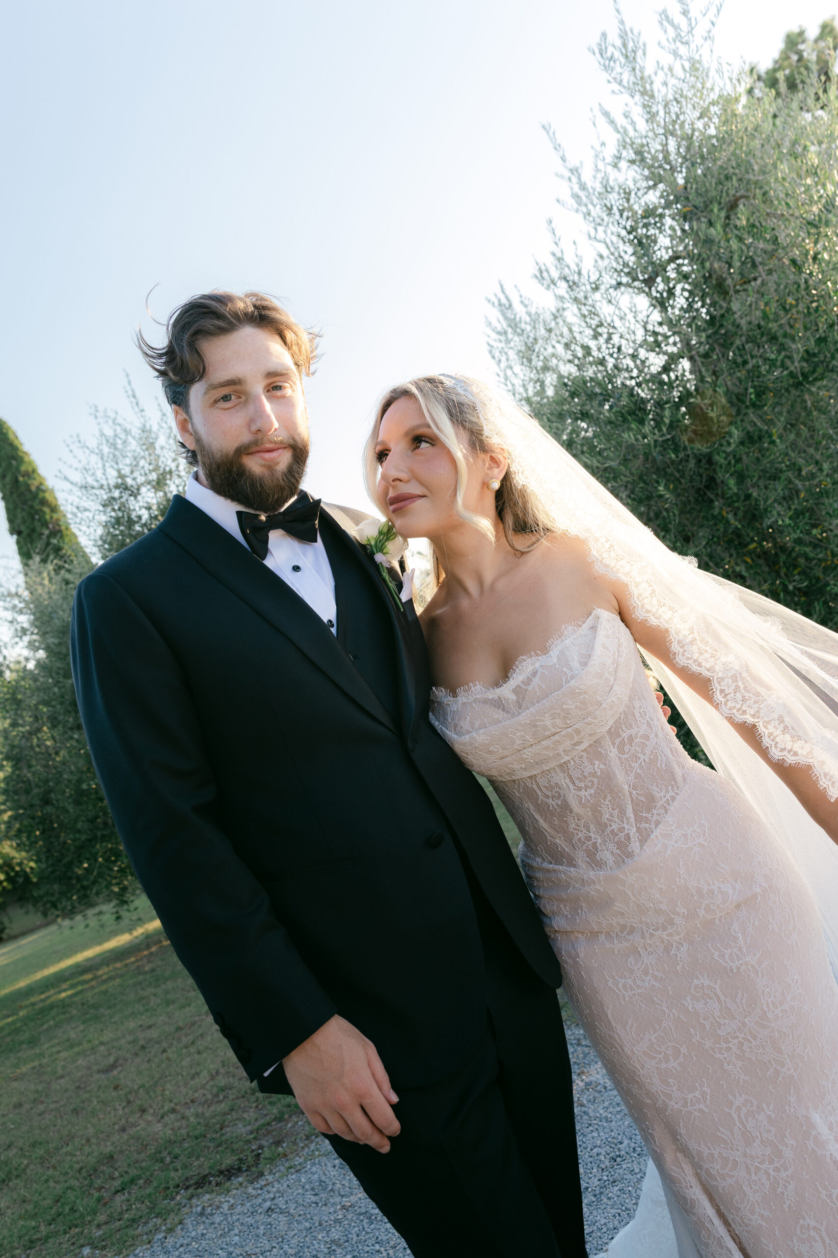 Bride in lace gown and long veil standing with groom in black tuxedo in the grounds of Villa Marchese Estate 1795 Tuscany