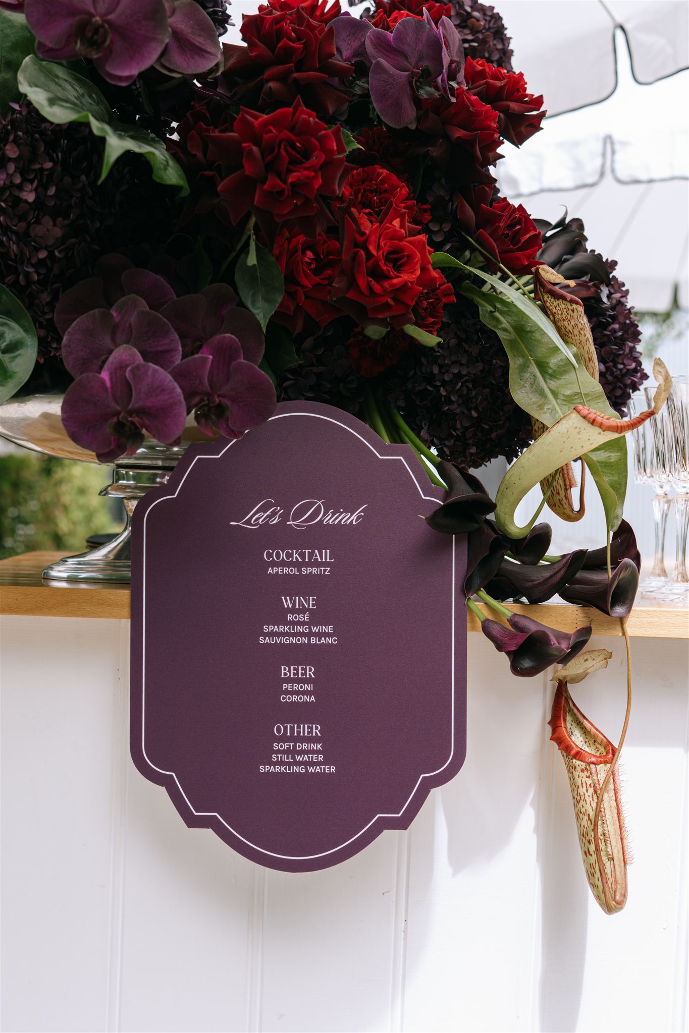 digital wedding photography, cocktail hour drinks menu sign surrounded by dramatic dark burgundy and deep red floral arrangement, sharper colour and detail