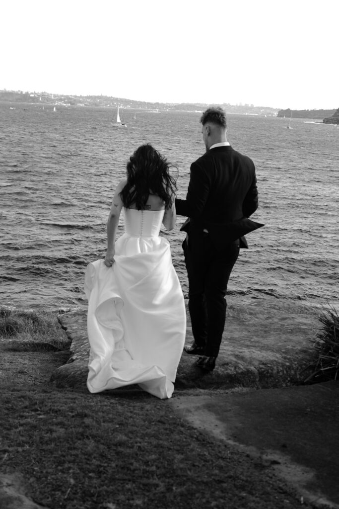Bride and groom walking hand in hand toward Manly Cove during wedding portraits