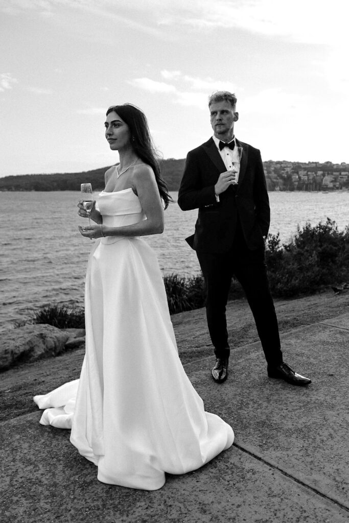 Bride in a full length gown and groom standing on the rocks along the Manly foreshore during wedding portraits