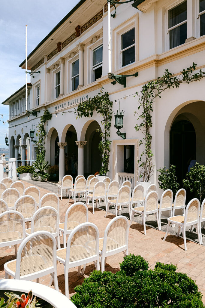Outdoor wedding ceremony setup at Manly Pavilion with rattan chairs and climbing florals on the heritage building