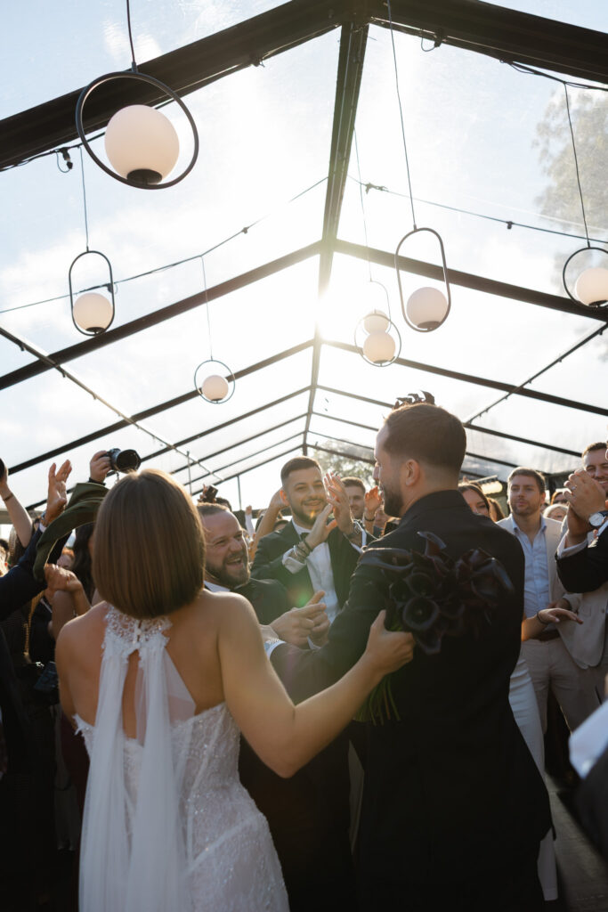 Bride and groom dancing with guests celebrating inside a clear marquee at Whites Valley Estate in McLaren Vale with sun flare.