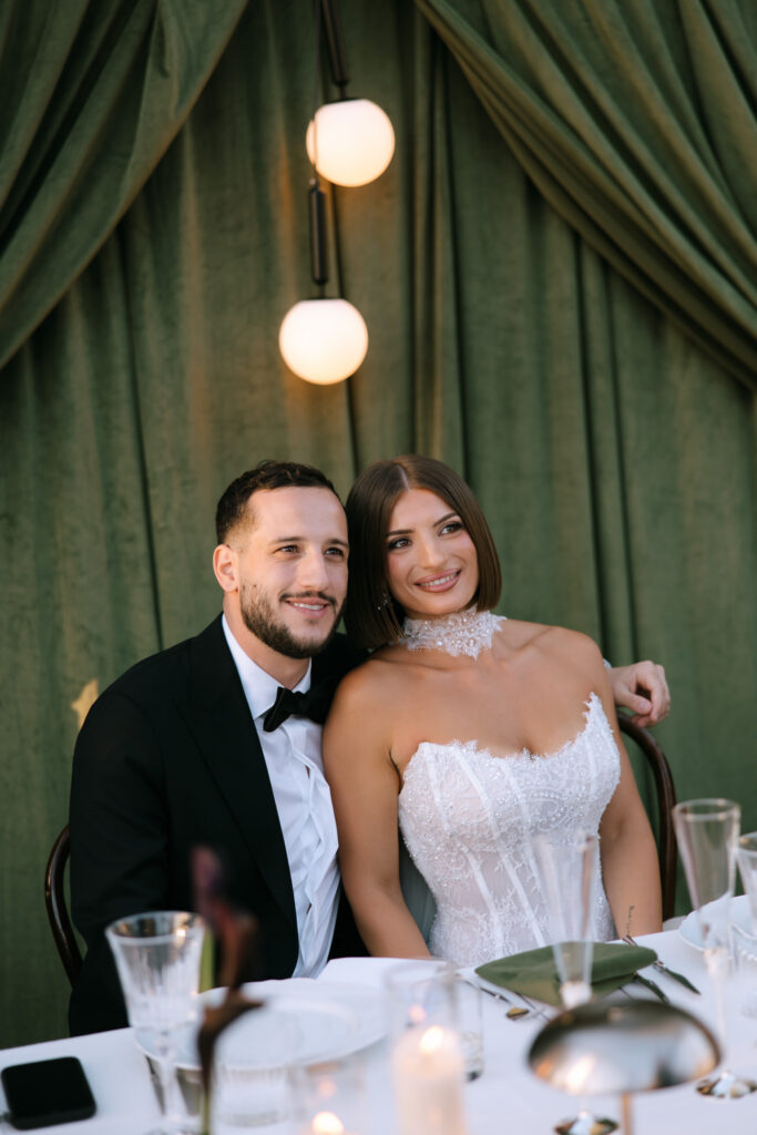 Bride and groom seated at the sweetheart table smiling against green velvet draping.