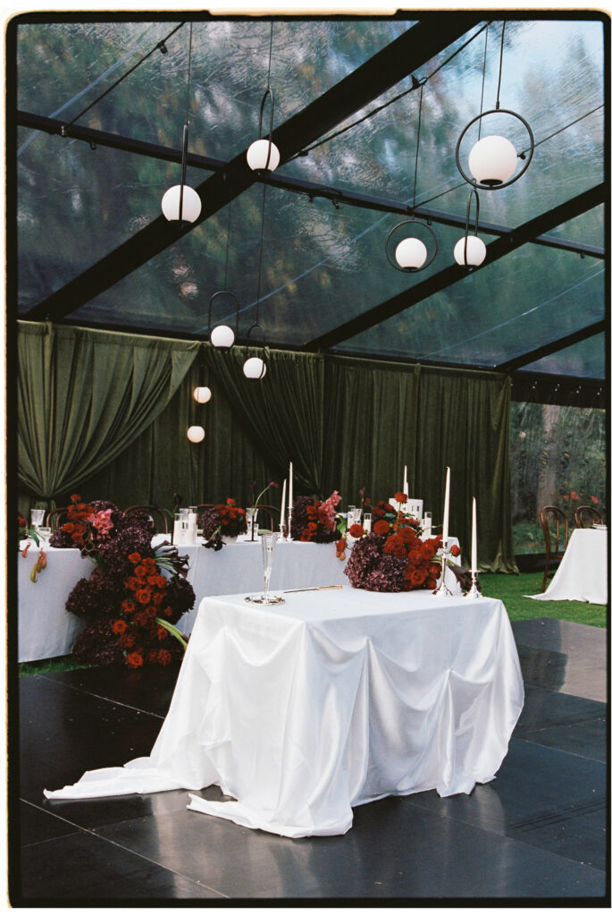 Sweetheart cake table with draped white linen, red and plum florals and pendant lights inside a clear marquee.