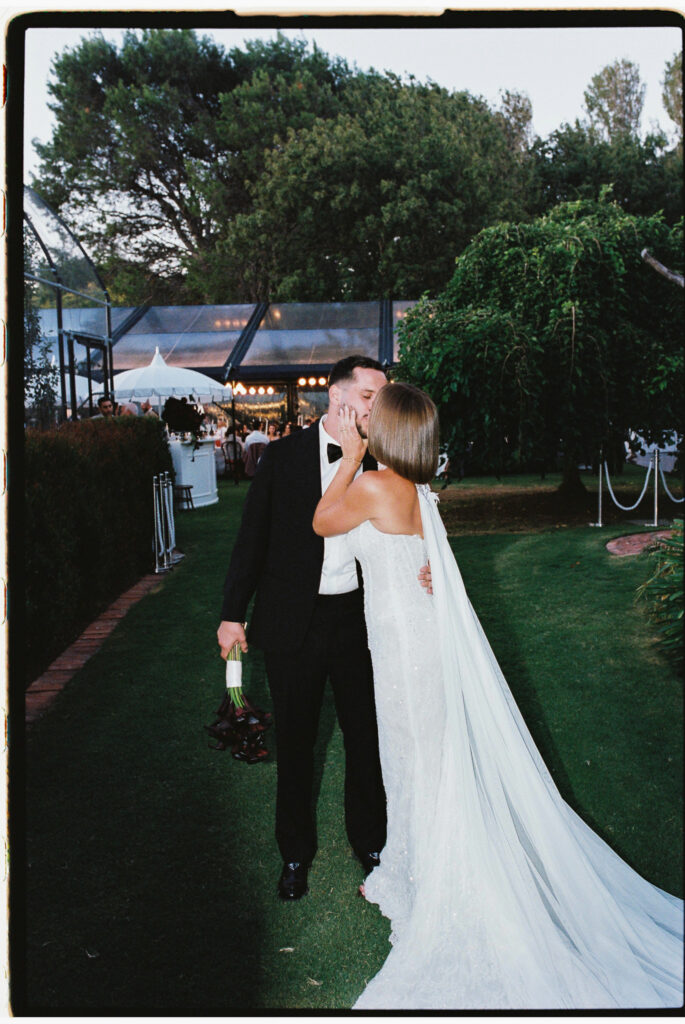 Bride and groom kissing on the lawn with the lit marquee and gardens of Whites Valley Estate in the background.