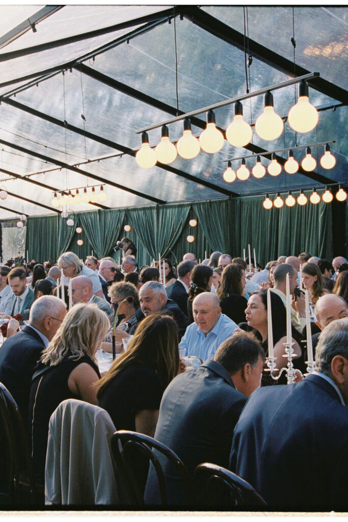 A 35mm film photograph of wedding guests seated at long tables inside a clear marquee with globe pendant lighting at Whites Valley Estate.