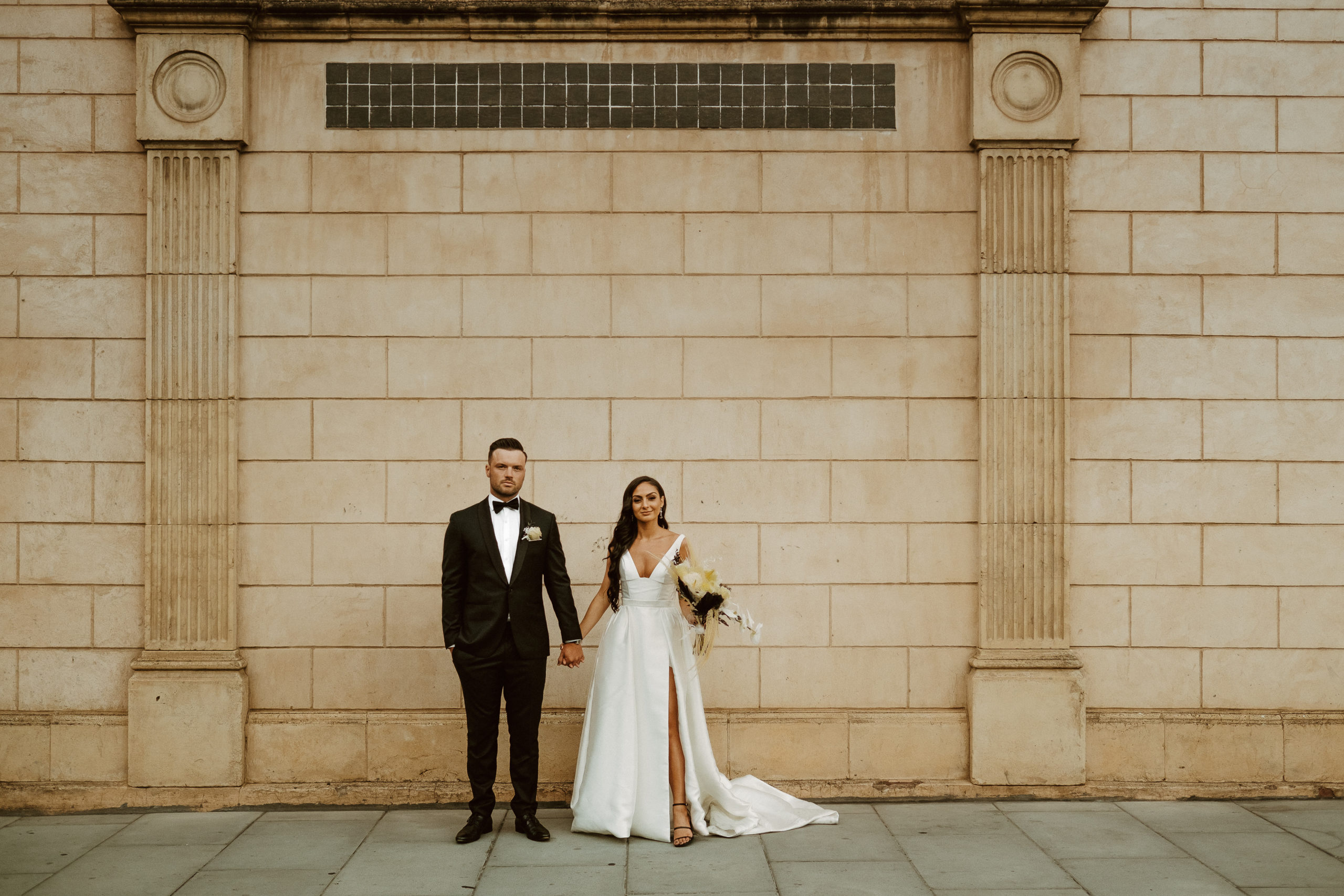 a modern bride and groom stand in front of a limestone building holding hands