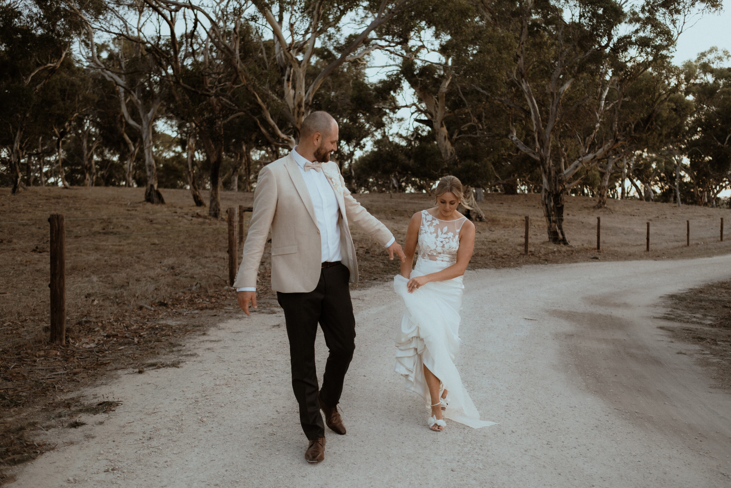 a newlywed couple walk down a winery driveway, the groom holding his brides hand, leading the way forward