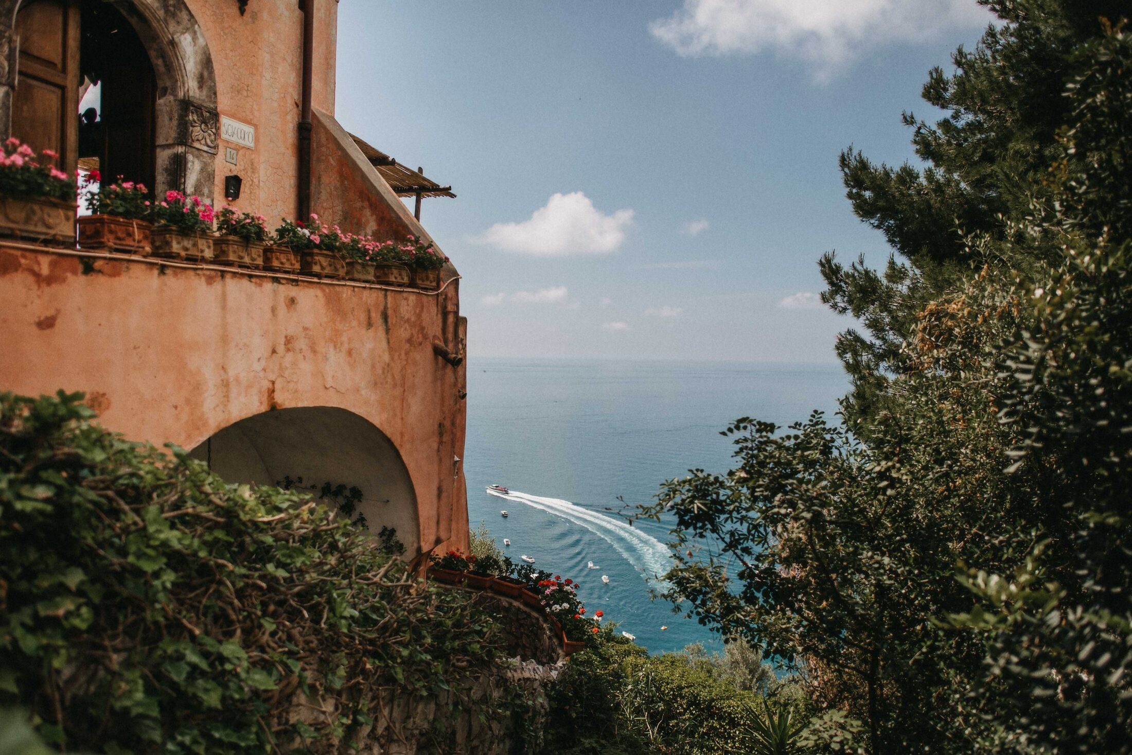 a view of the ocean from Positano, a yacht speed away on the water. a Tuscan villa with open archways sits on the left. a green tree to the right, framing the ocean view. destination wedding.