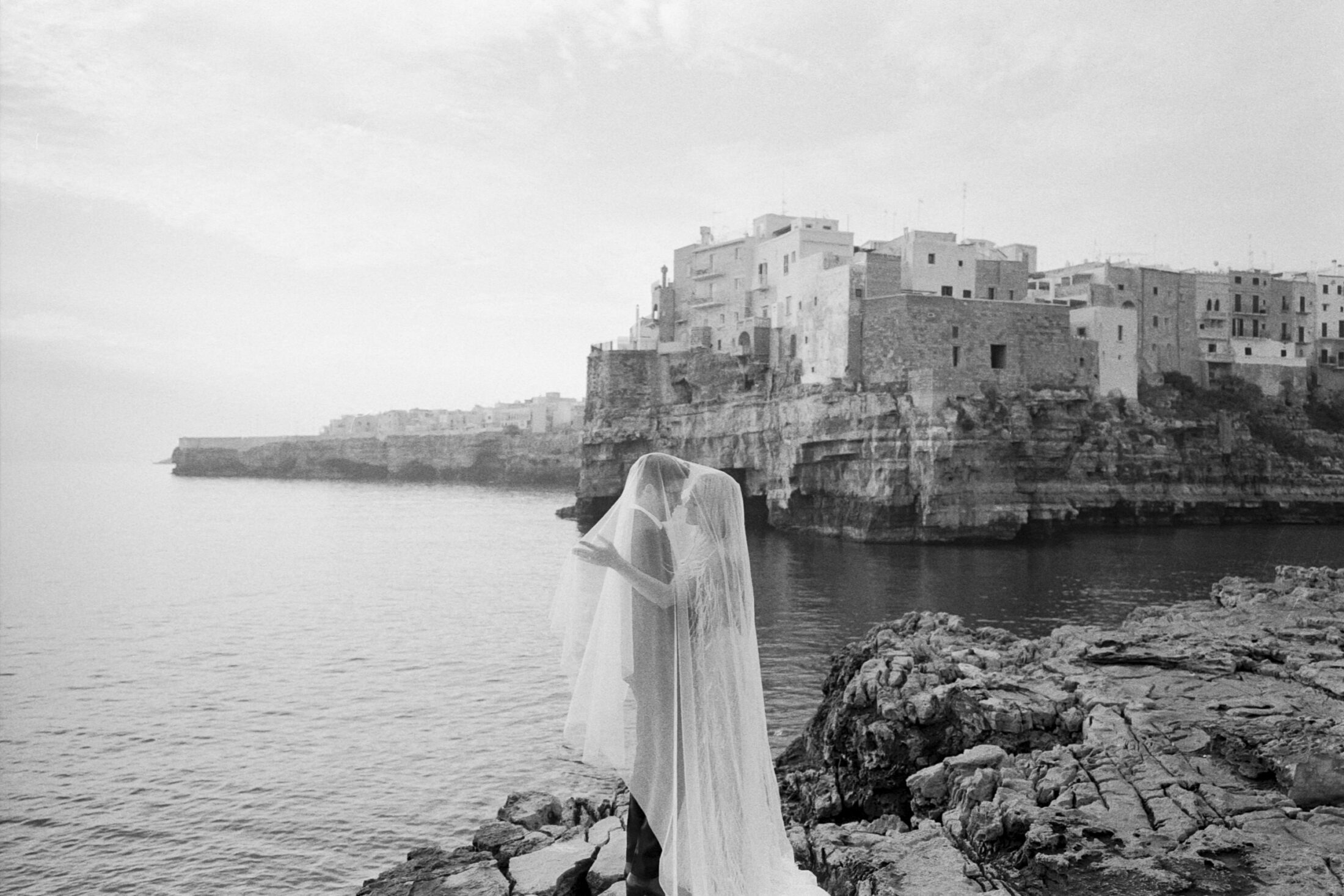 a bride and groom embrace under the bride's veil along a rocky coast in Puglia, Italy. destination wedding.