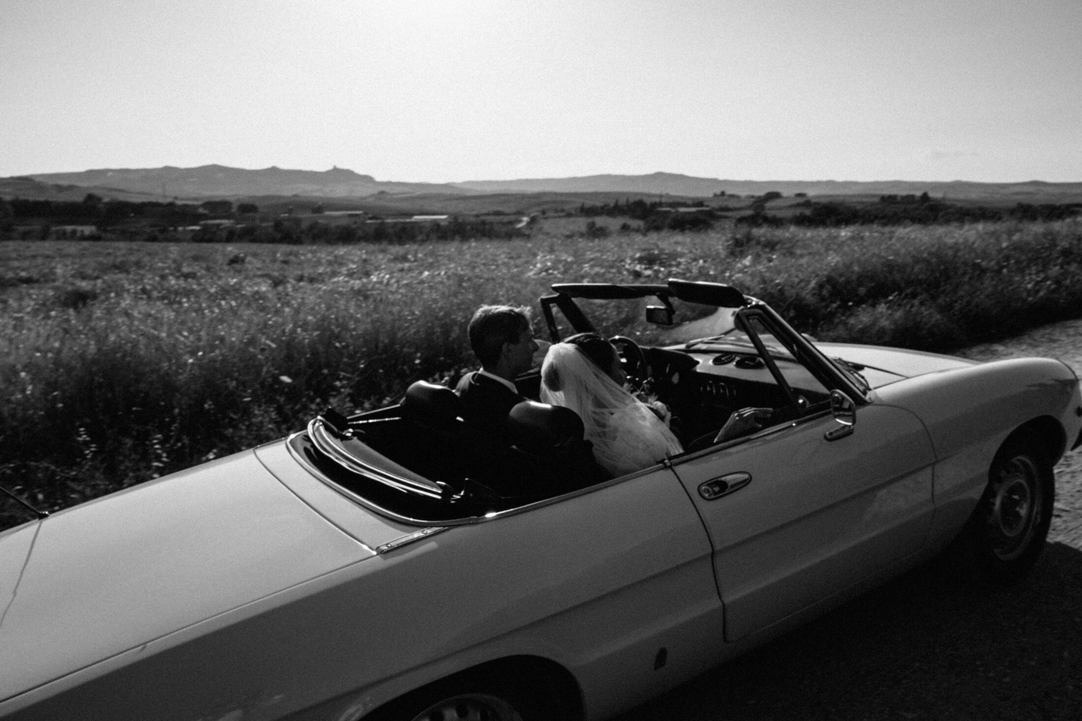 a black and white photograph of a bride and groom in an old convertible driving through the Italian countryside during a destination wedding.
