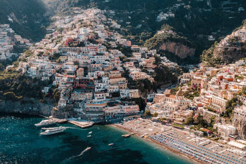 an ariel photograph of the Positano coastline. a beach filled with blue umbrellas, surrounded by a village built into the sunlit hills and clifftops. yachts are docked at the small jetty.