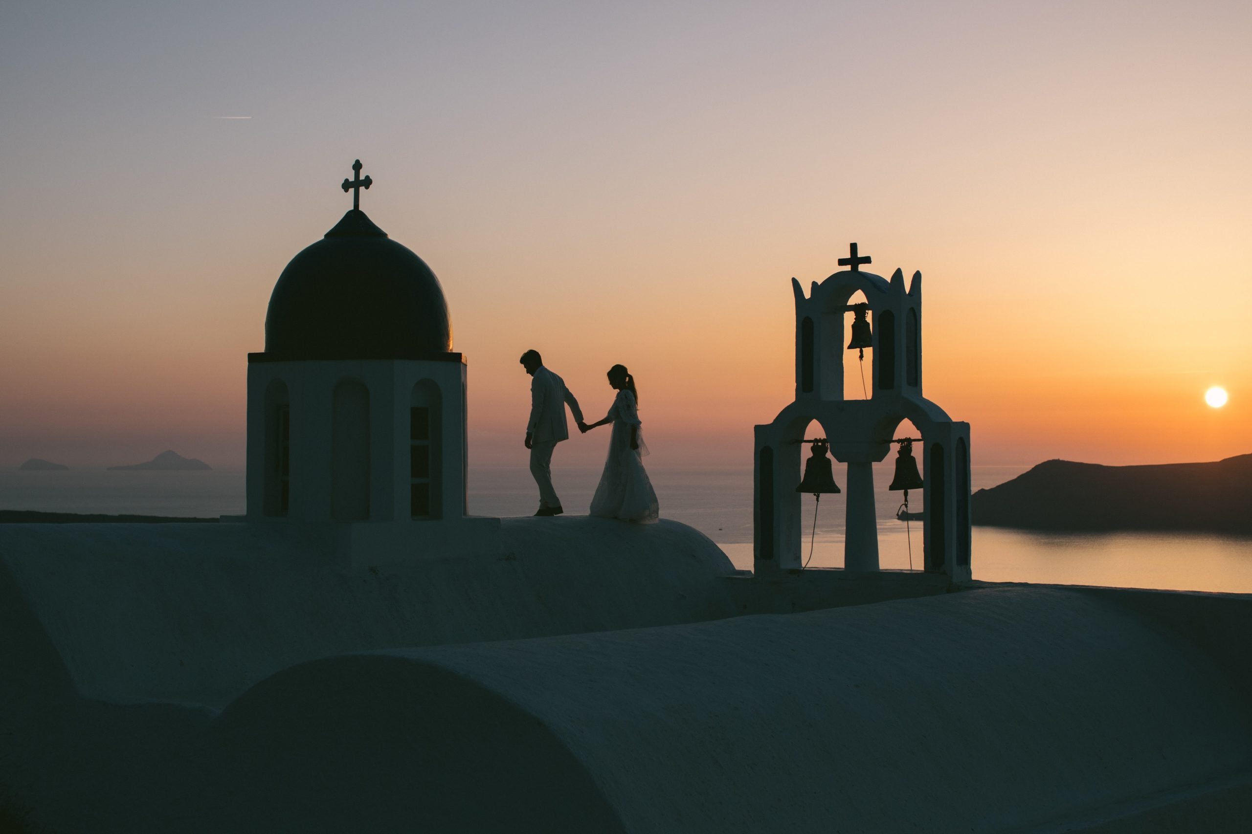 a newlywed couple walking on a rooftop in Santorini at sunset. the groom holds his brides hand, leading her. destination wedding.