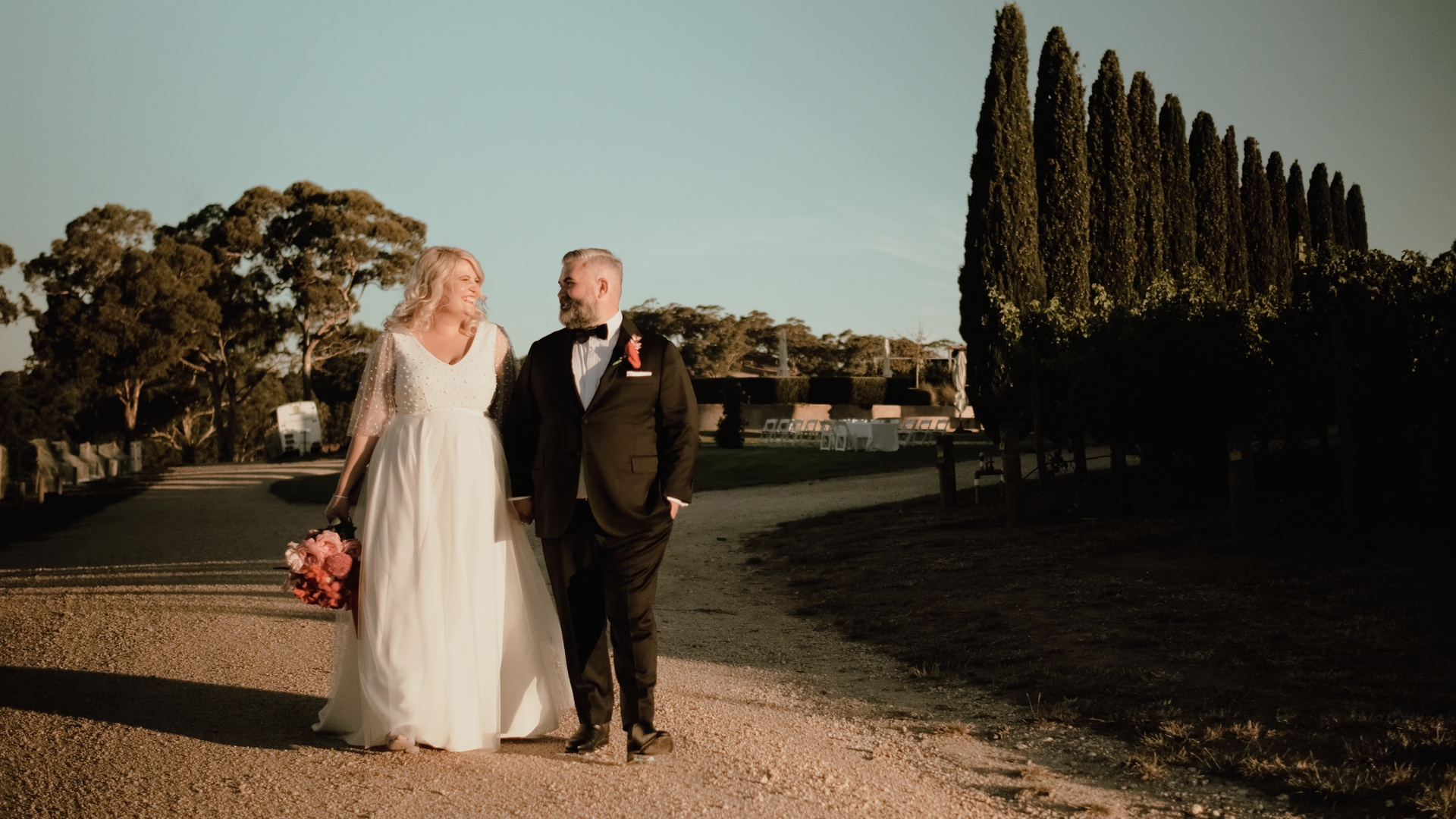a bride and groom stroll down a driveway together at sunset, cyprus trees to their right at The Lane Vineyard