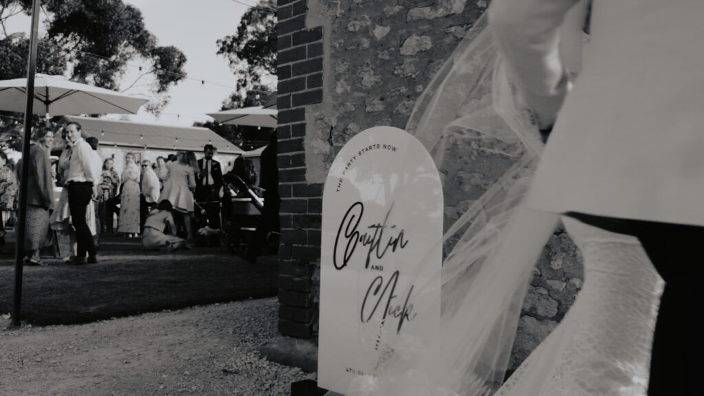 a black and white image of a bride and groom walk past their wedding signage into the stone outdoor chapel at Woodburn Homestead