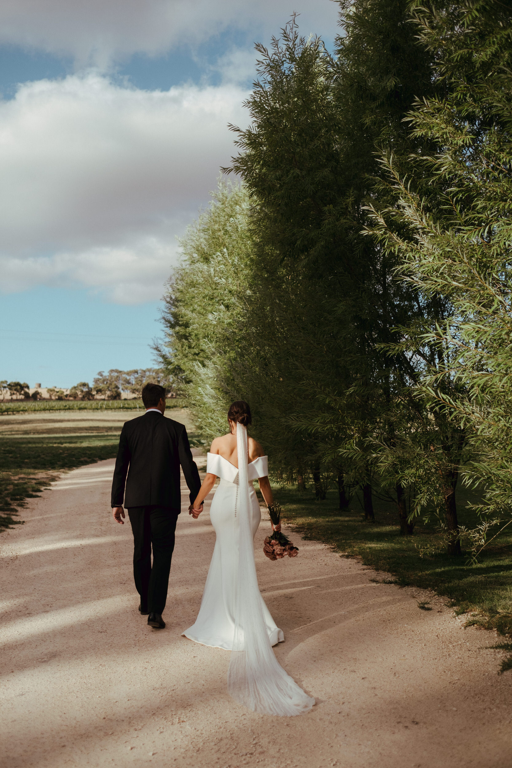 a bride and groom walk down a tree-lined driveway at Howard's
