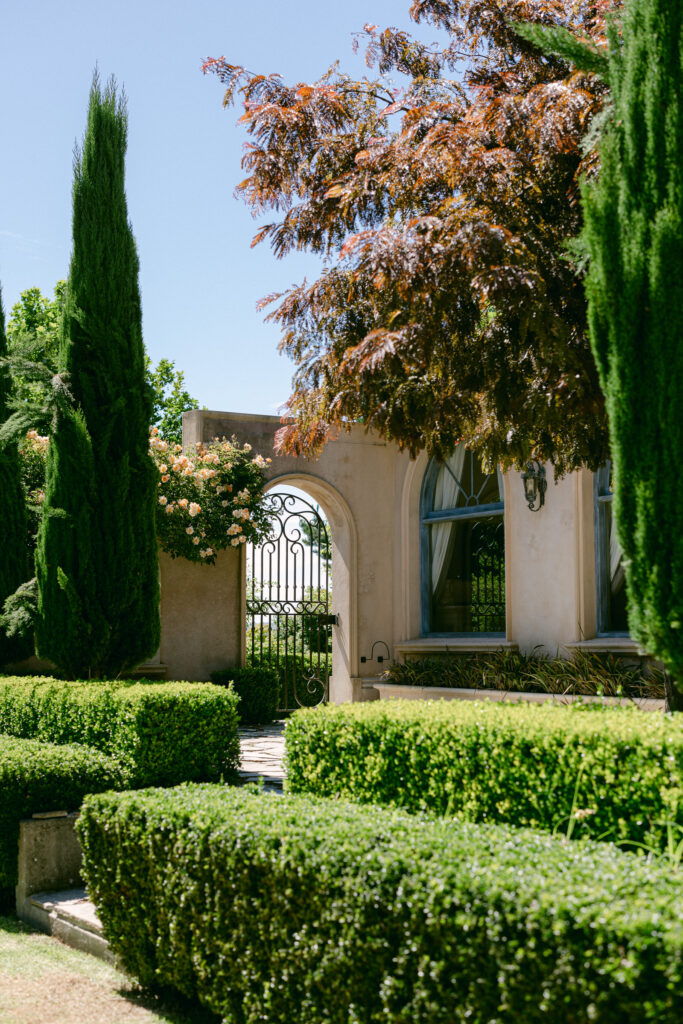 Manicured hedges and tall cypress trees framing the stone archway and iron gate at Villarica, a Tuscan-inspired wedding venue in the Adelaide Hills, South Australia.