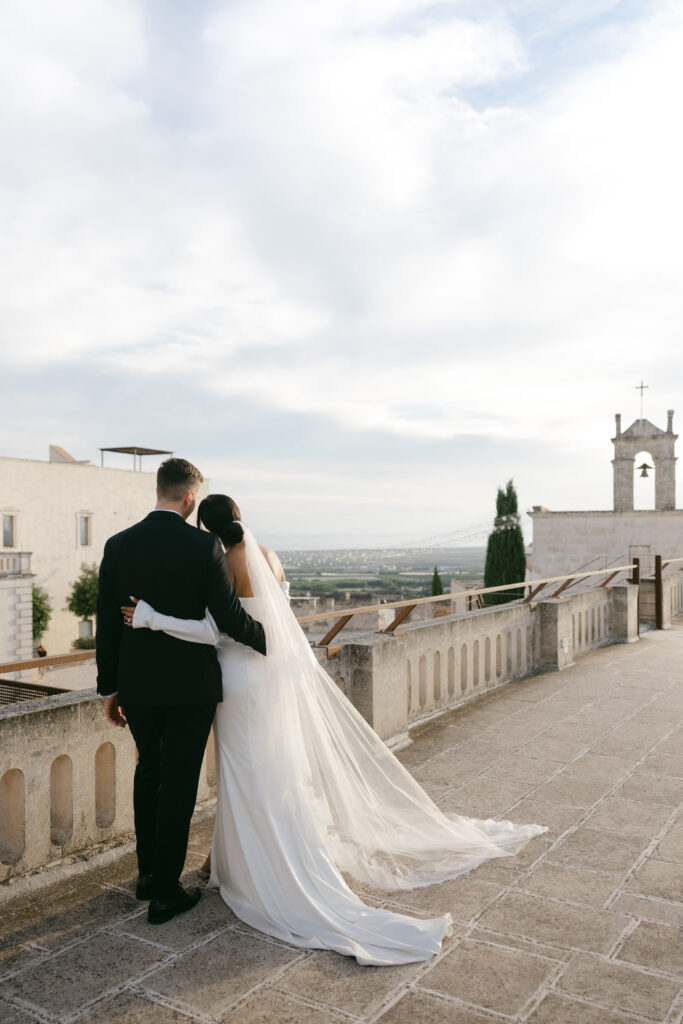 Bride and groom hugging along the rooftop terrace at Masseria Amastuola in Puglia, admiring their wedding reception in the courtyard below