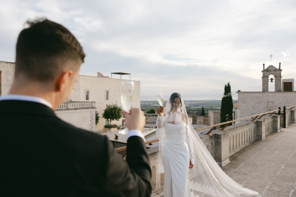 Bride toasting a glass of champagne to her groom with the courtyard of Masseria Amastuola in Puglia in the background