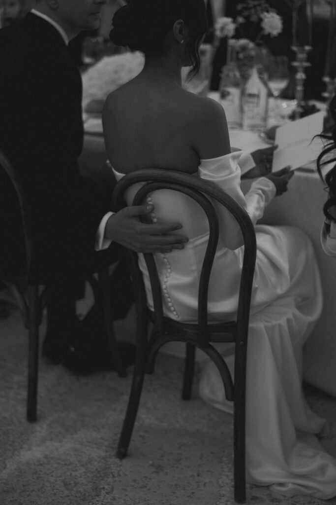 Black and white moment of groom's hand placed on the back of bride's chair during their wedding dinner at Masseria Amastuola