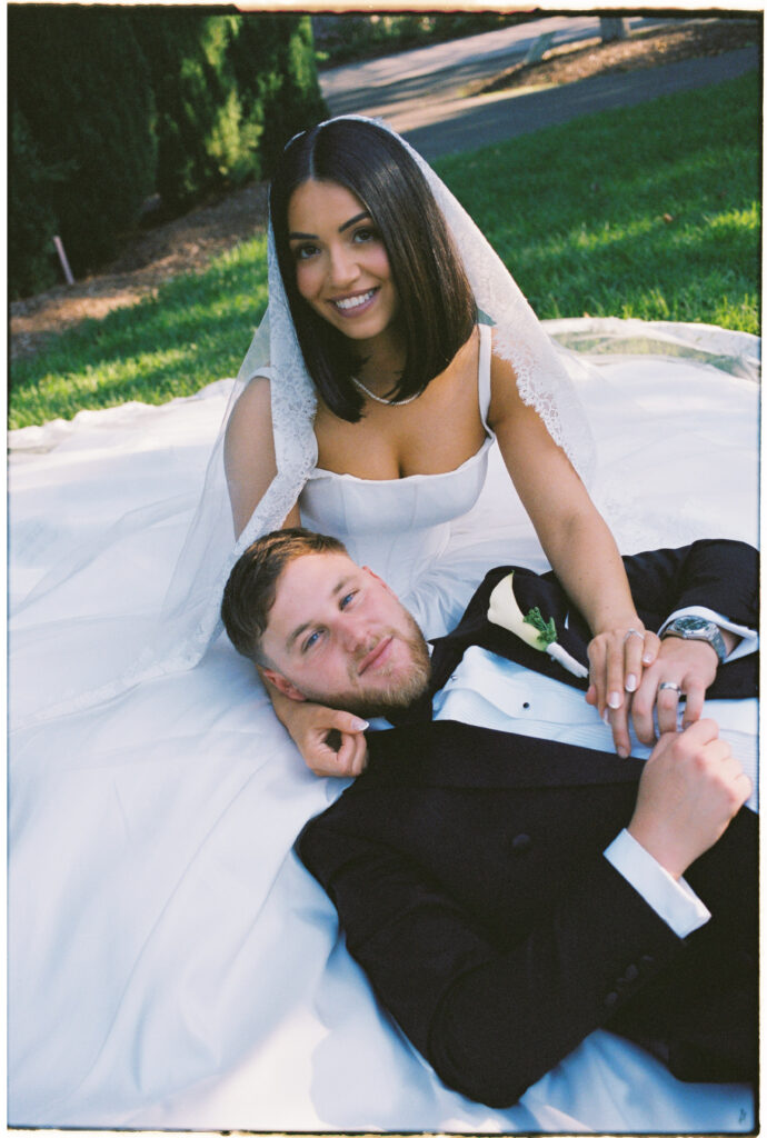 35mm film wedding photography, bride and groom lying on grass laughing together, bride in square neck gown with long veil draped over groom
