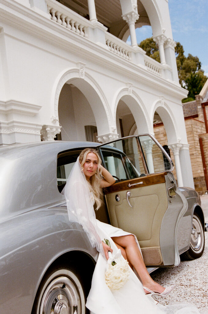 35mm film wedding photography, bride in strapless gown sitting in vintage cream car doorway with veil cascading down, heritage building in background
