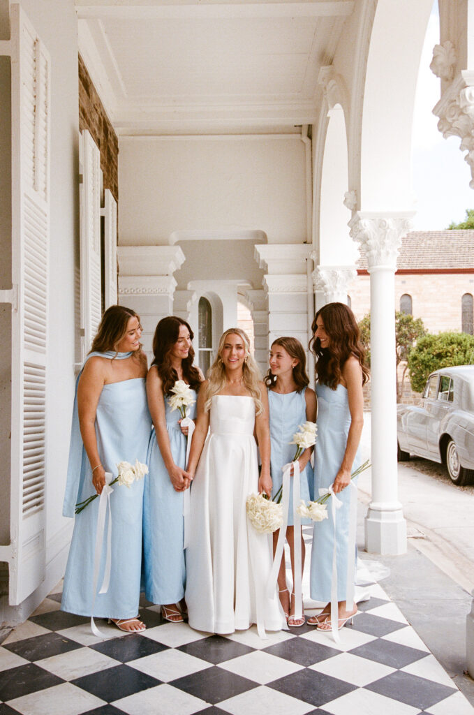 35mm film wedding photography, bride with four bridesmaids in powder blue gowns standing on verandah of heritage white colonial building