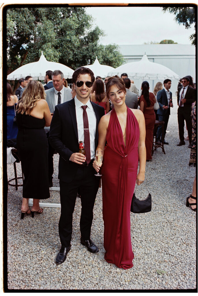 35mm film wedding photography, couple at outdoor cocktail hour, woman in deep red halter gown and man in dark suit holding drinks, marquee in background
