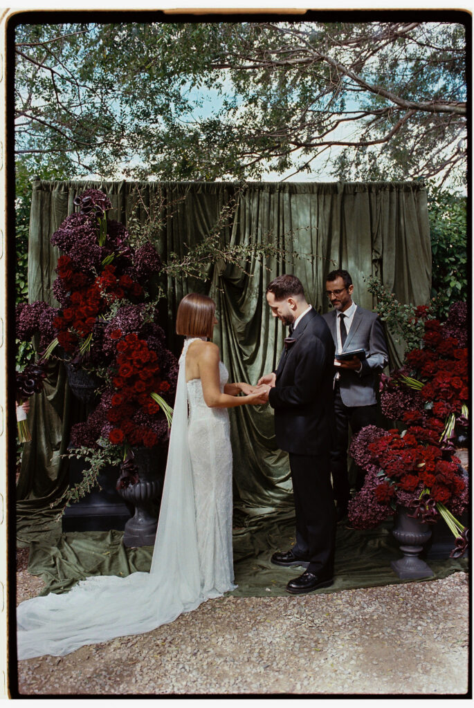 35mm film wedding photography, bride and groom exchanging vows at outdoor ceremony beneath dramatic floral arch with dark red florals and sage green draping, celebrant present
