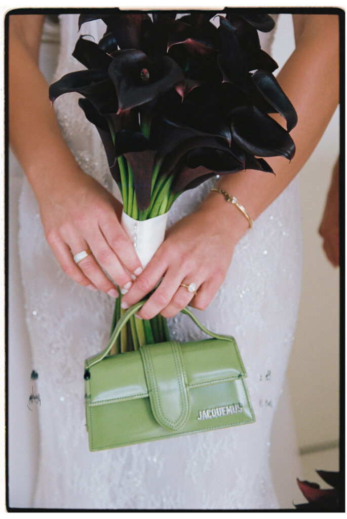 35mm film wedding photography, close up of bride's hands holding dramatic black calla lily bouquet with small green Jacquemus bag at wrist, sequin gown detail
