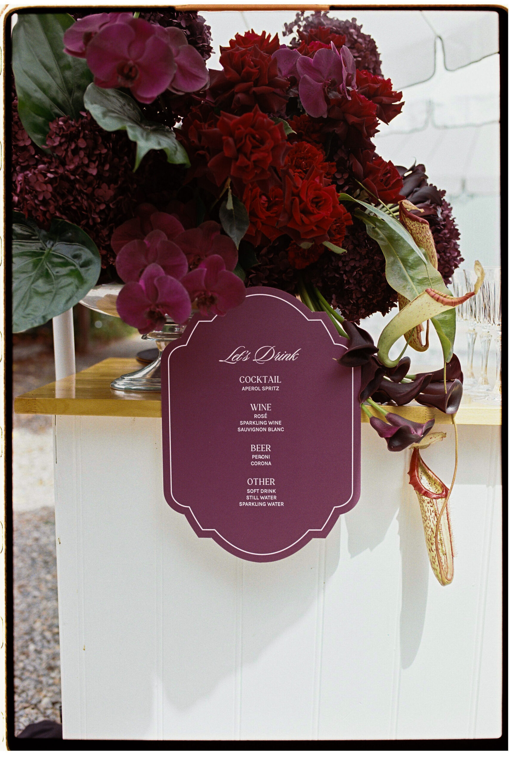 35mm film wedding photography, cocktail hour drinks menu sign surrounded by dramatic dark burgundy and deep red floral arrangement
