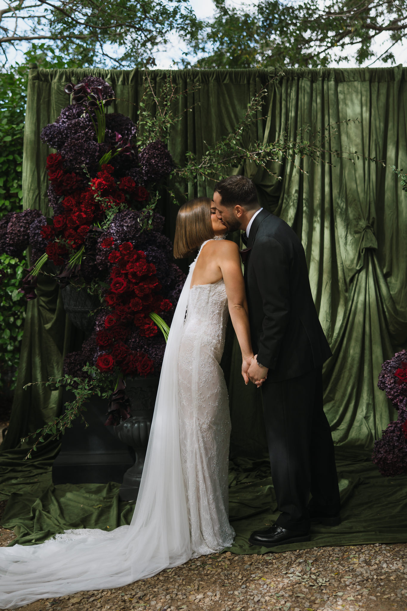 Bride and groom sharing a kiss in front of a lush floral sculptural installation with dark green drapery at Whites Valley Estate.