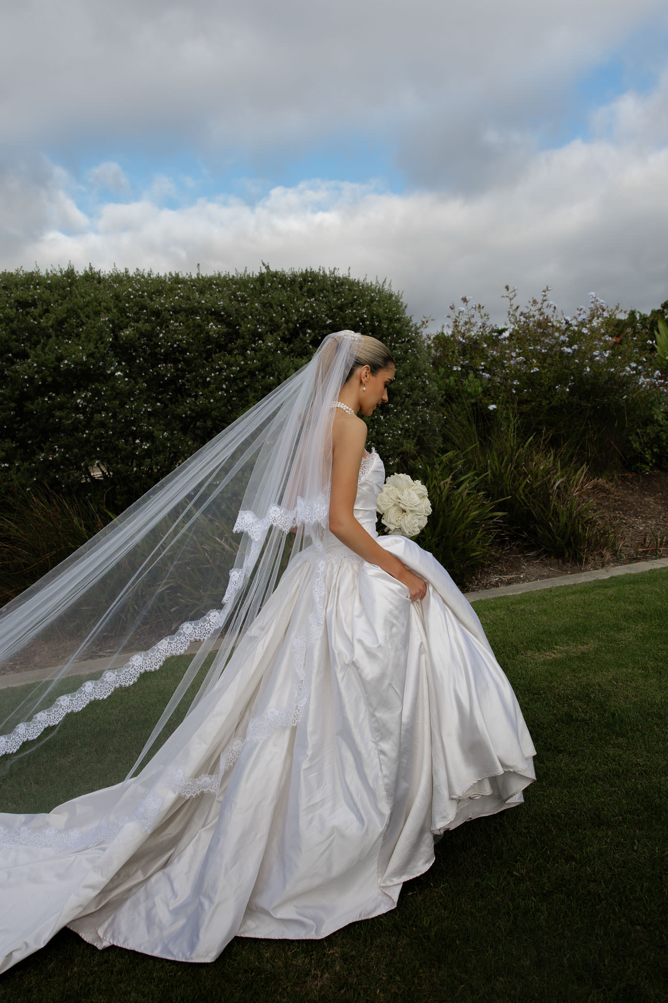 Bride walking across the lawn at Beresford Estate in a full cathedral veil, hedging framing the background.